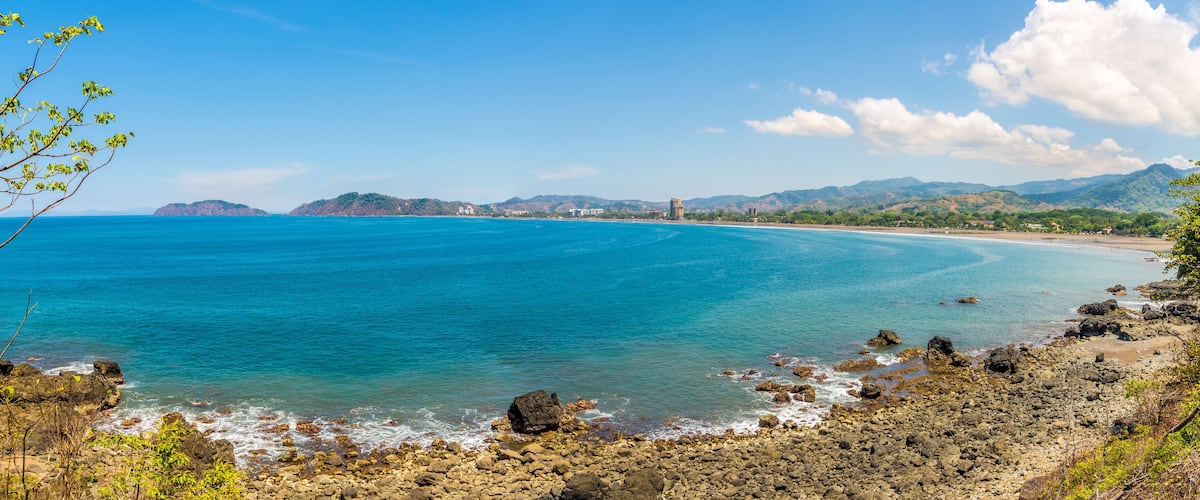 A coastal landscape with clear blue waters, rocky shore, and distant mountains.