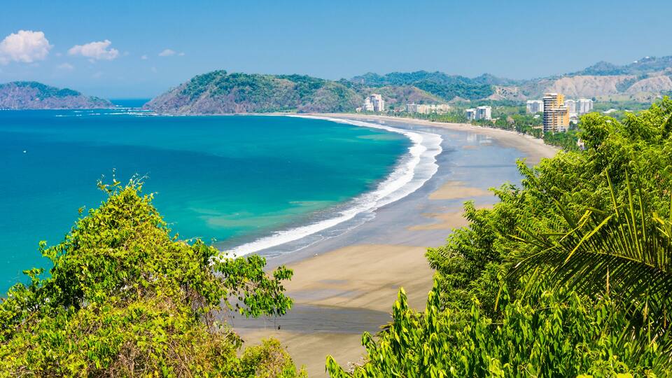 A coastal landscape with a sandy beach, clear turquoise waters, and lush green hills in the background.