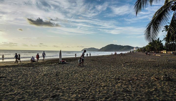 A beach with people walking, a surfboard, palm trees, and a distant mountain.