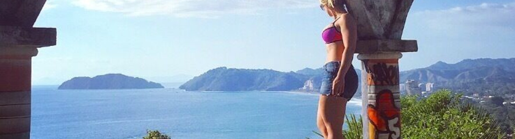 Uma mulher está em uma varanda com vista para a costa, sob um céu claro.