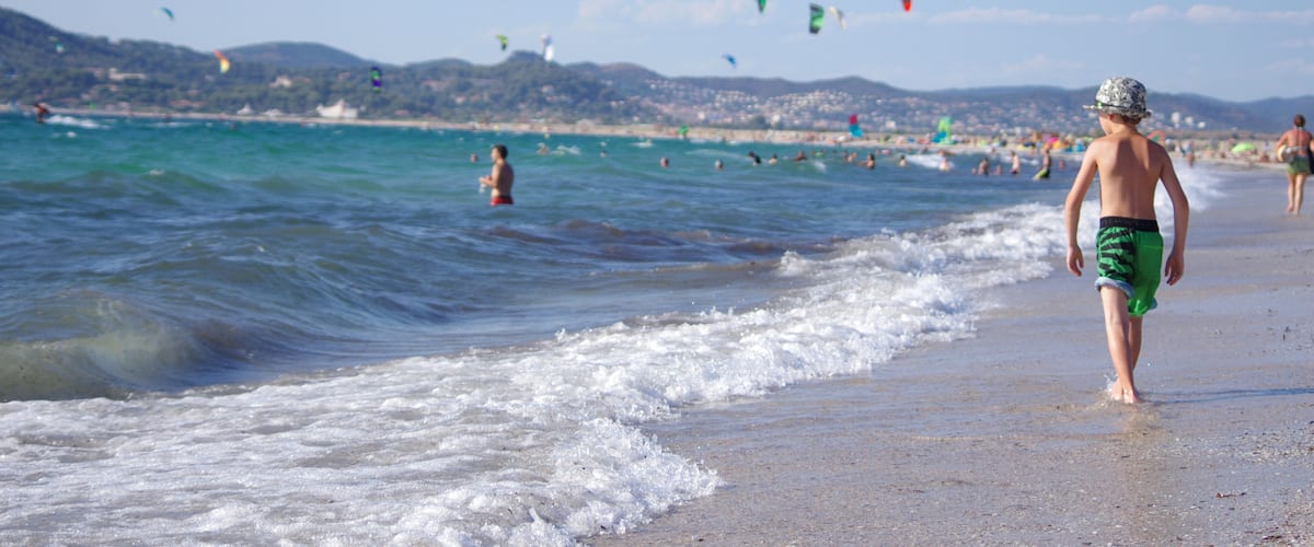 A beach with people swimming and walking along the shore, and colorful kites flying in the sky.