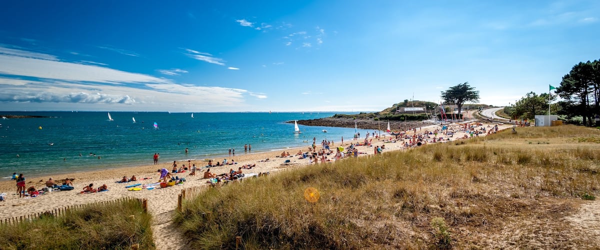 A beach with people sunbathing, a sandy shore, and sailboats in the water.