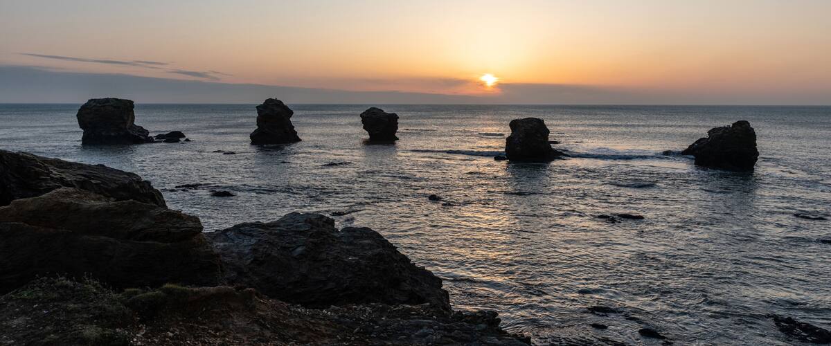 A sunset over a calm sea with prominent rock formations in the foreground.