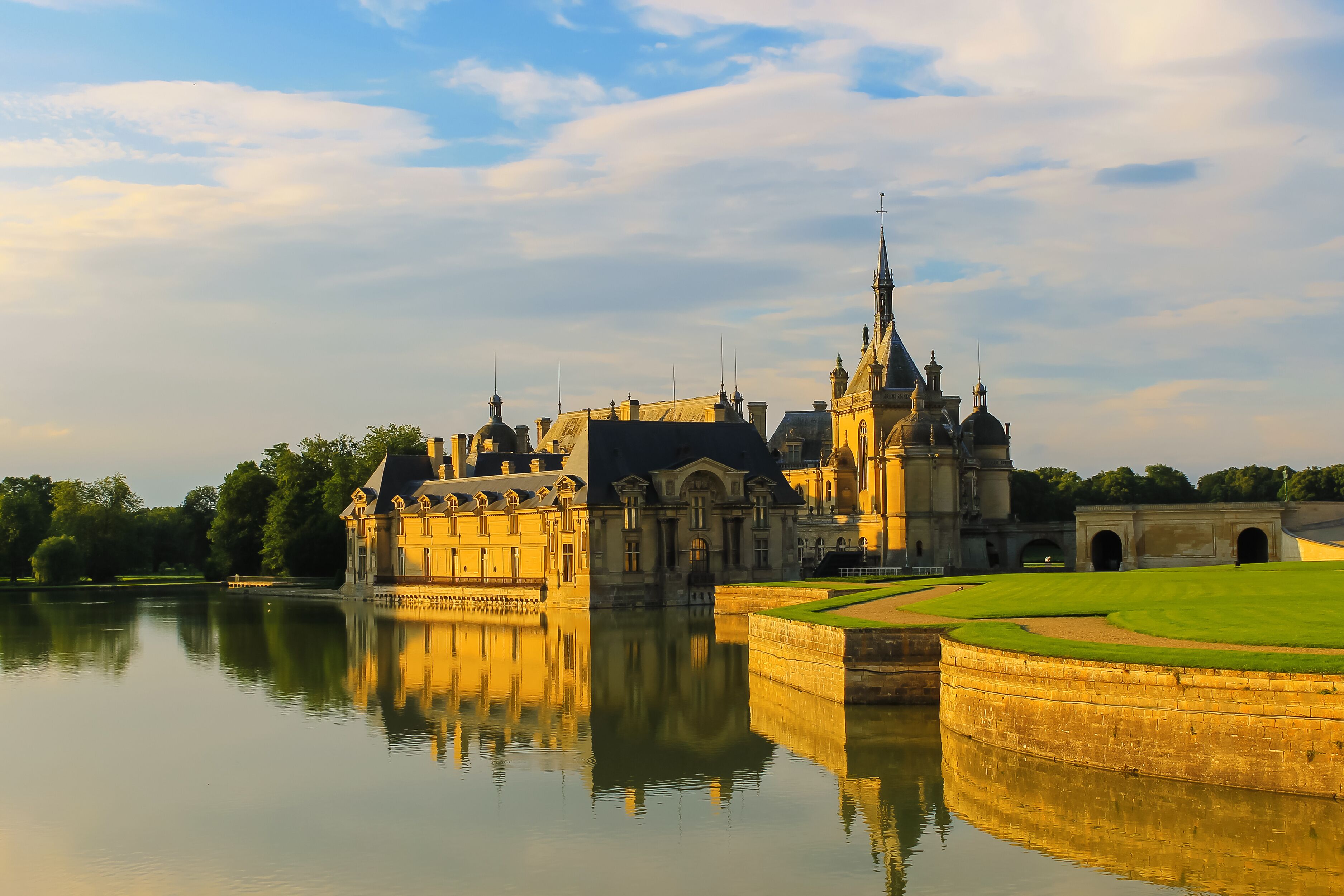 A grand castle with a moat, surrounded by lush greenery and a calm lake reflecting its architecture.