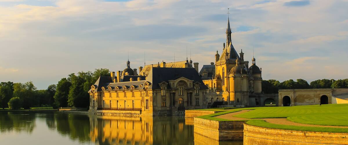 A grand castle with a moat, surrounded by lush greenery and a calm lake reflecting its architecture.