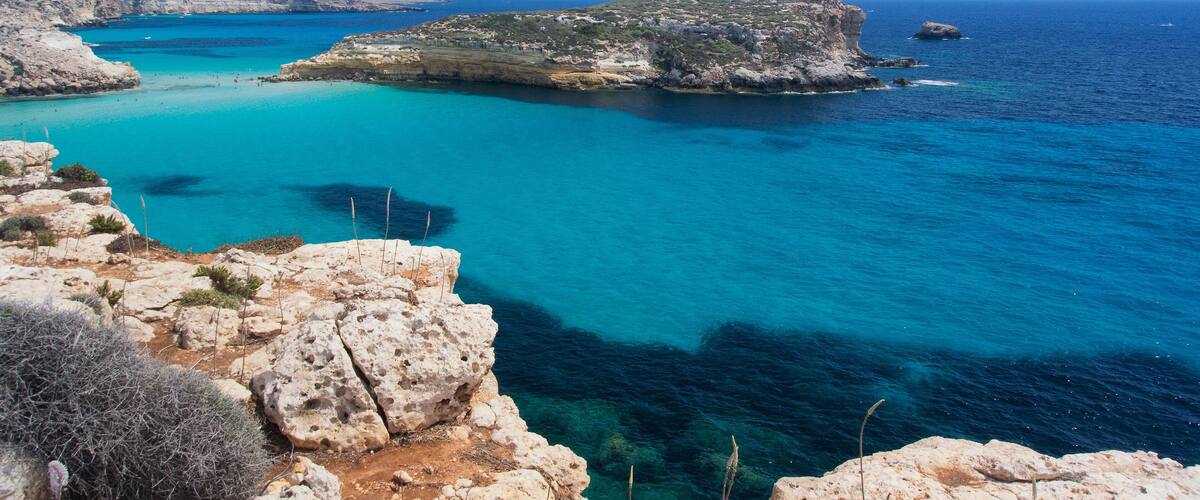 A coastal landscape with clear turquoise waters, rocky cliffs, and sparse vegetation.
