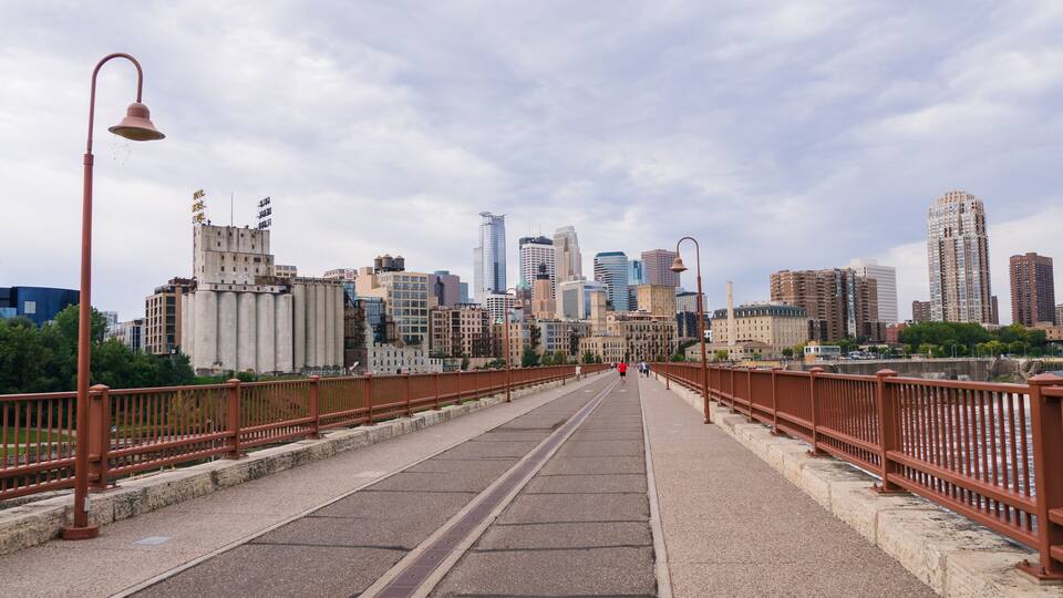 A pedestrian bridge with a metal railing, leading towards a city skyline with various buildings.