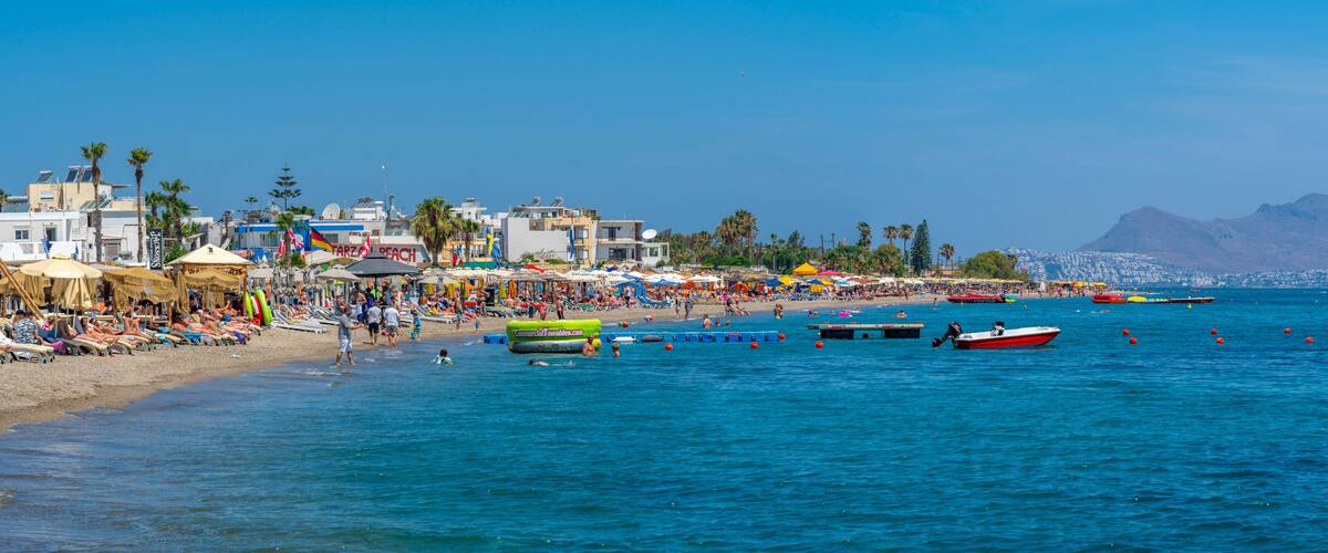 A crowded beach with people sunbathing, umbrellas, and boats.
