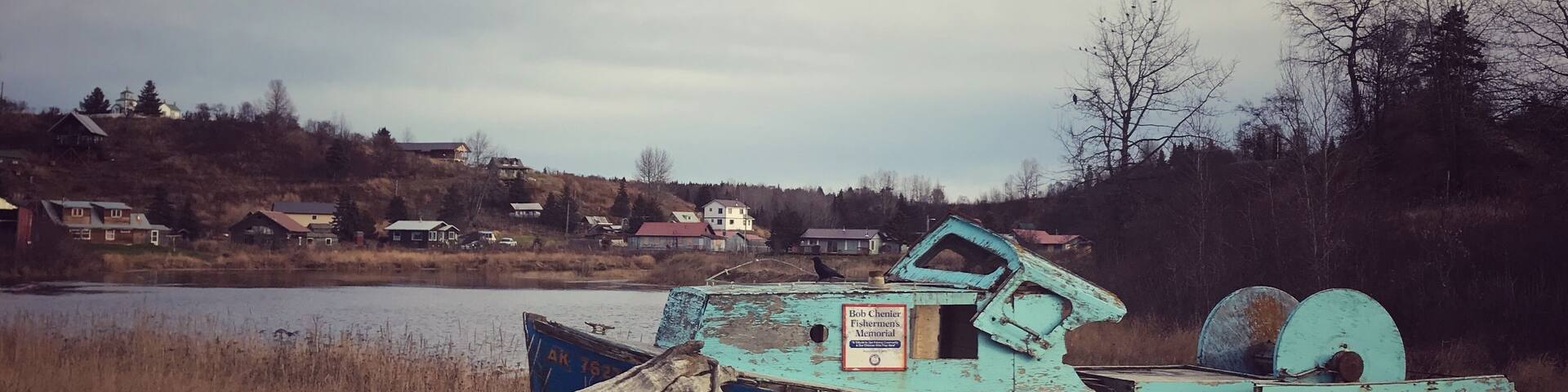 Un bateau bleu, sur lequel on peut lire l’inscription « Mémorial des pêcheurs Bob Chenier », est amarré au bord d’un lac, avec des maisons en arrière-plan.