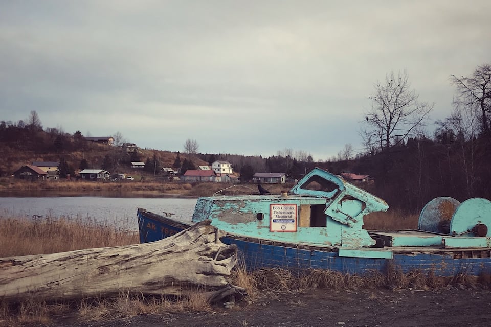 A blue boat with a sign labeled "Bob Chenier Fishermen's Memorial" by a lake with houses in the background.