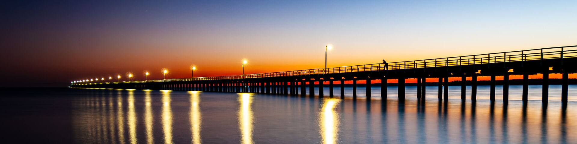 A long pier extending into the water at sunset with lights reflecting on the surface.