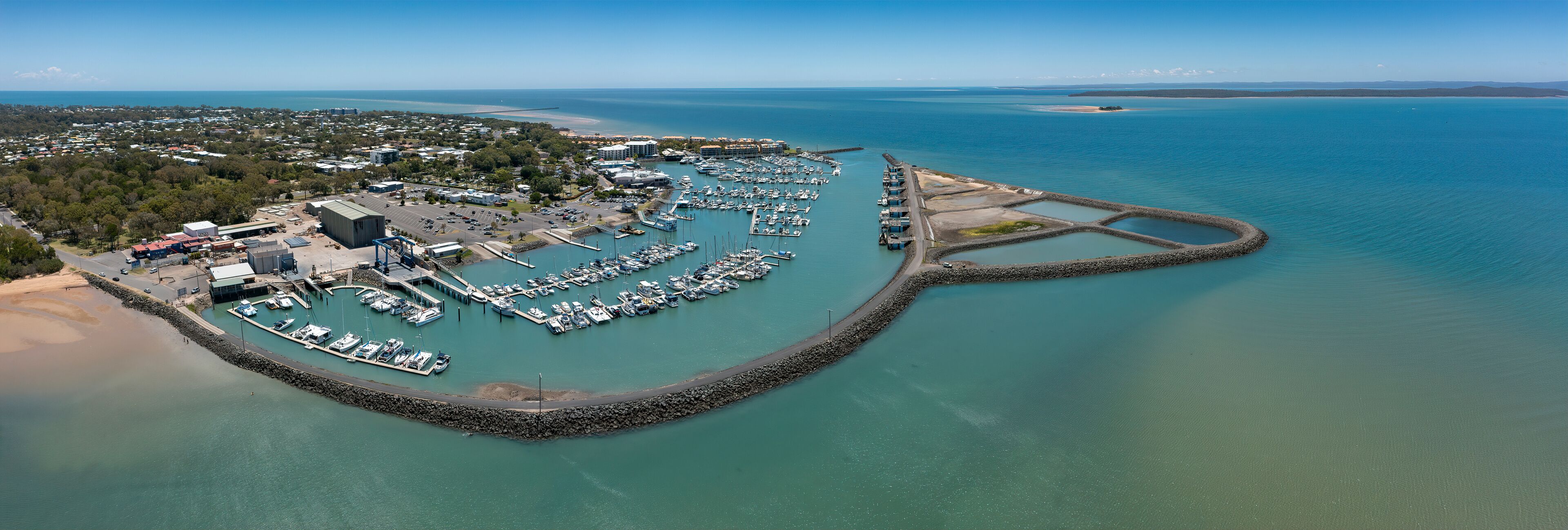 Vista aérea de un puerto deportivo costero con barcos amarrados, rodeado por un rompeolas rocoso y una zona boscosa.