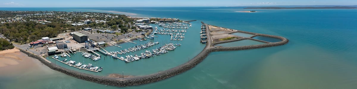 Vue aérienne d’un port de plaisance situé sur la côte, avec des bateaux amarrés, entouré d’une digue rocheuse et d’une zone boisée.