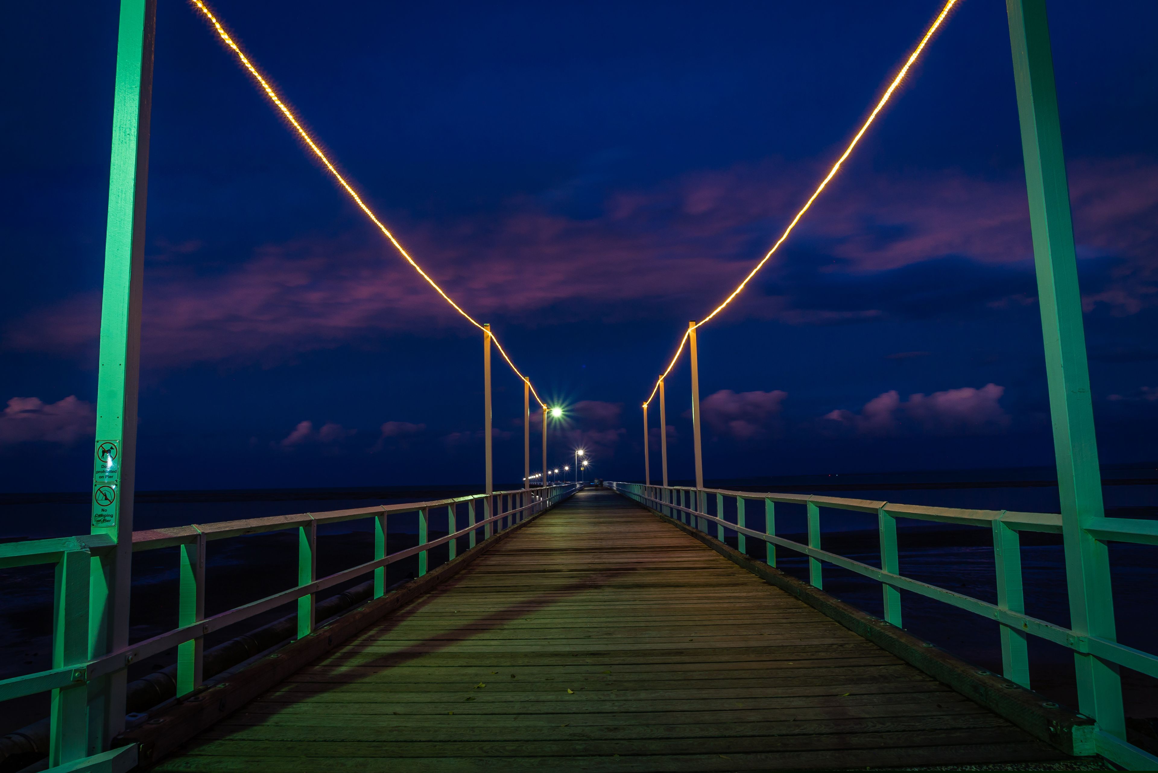 A wooden pier with green railings and lights, extending into the night.