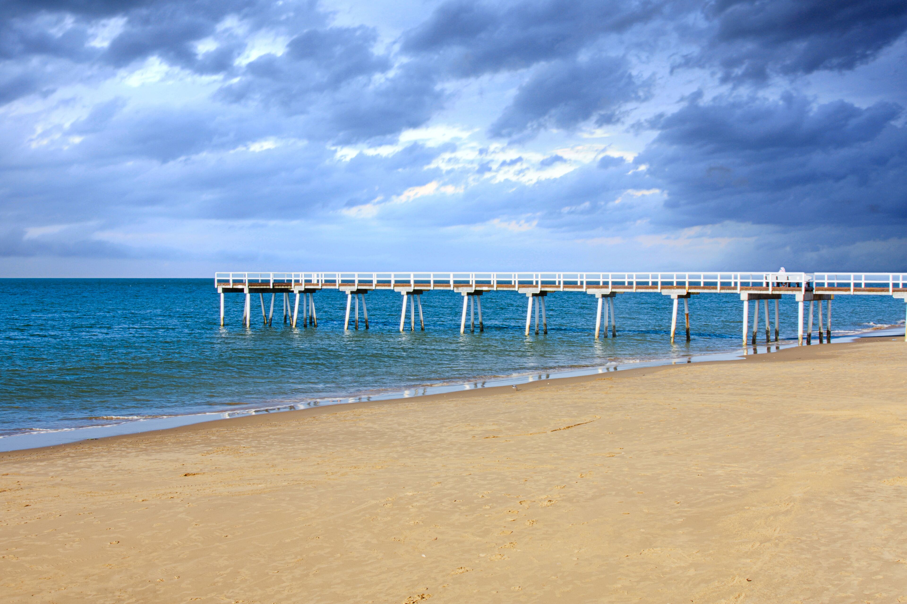 A beach with a pier extending into the ocean under a cloudy sky.