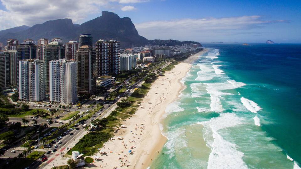 Un paisaje urbano costero con rascacielos, una playa de arena y una montaña al fondo.
