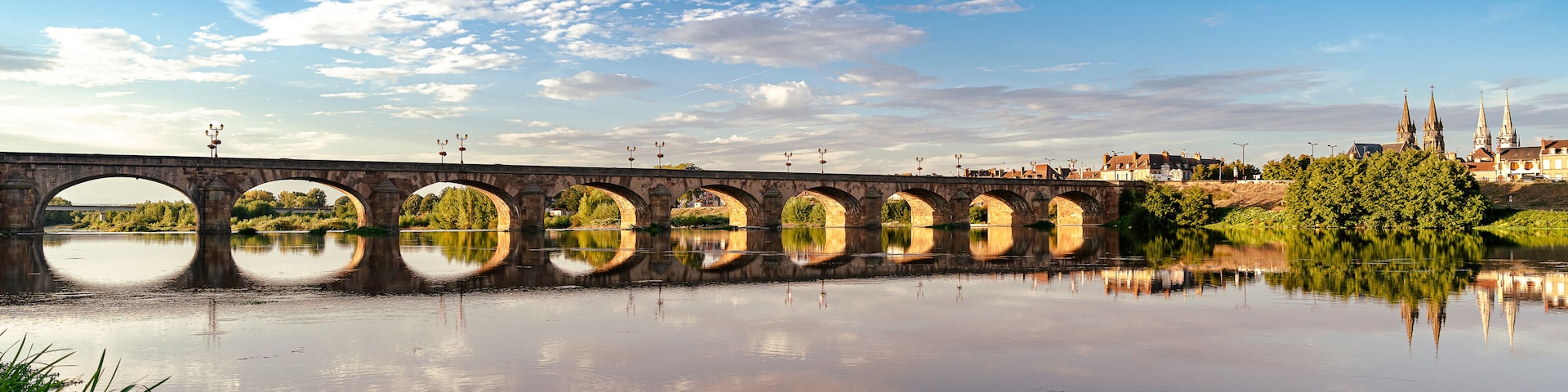 Un puente de piedra con múltiples arcos sobre un río tranquilo, rodeado de vegetación y un cielo azul despejado.