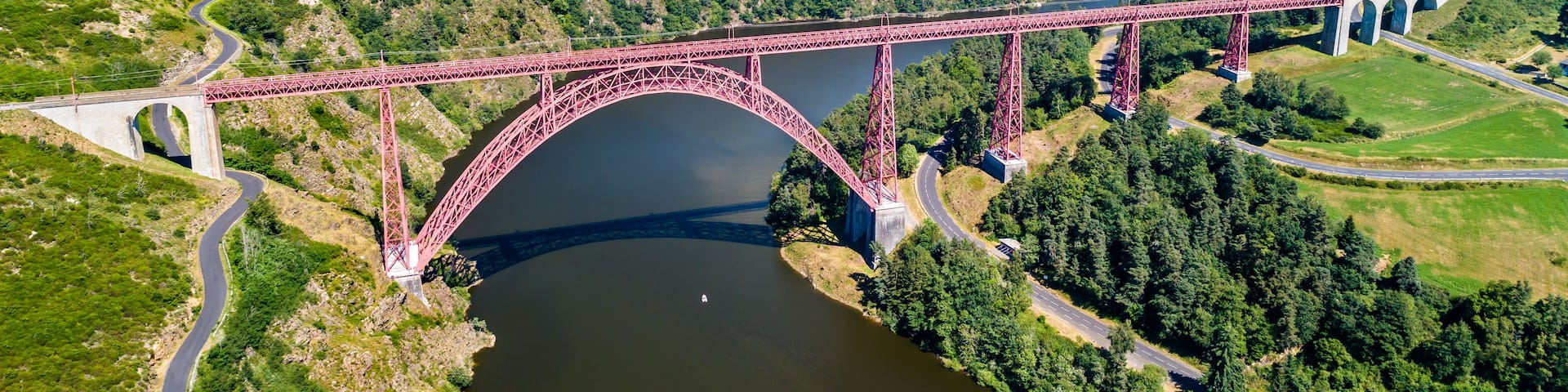 A red steel bridge spanning a river, surrounded by lush greenery and rolling hills.