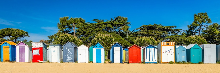 A row of colorful beach huts with a clear blue sky and trees in the background.