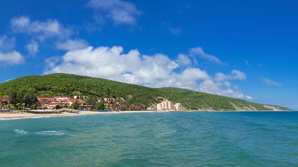 A coastal landscape with a beach, a mountain, and buildings along the shore.