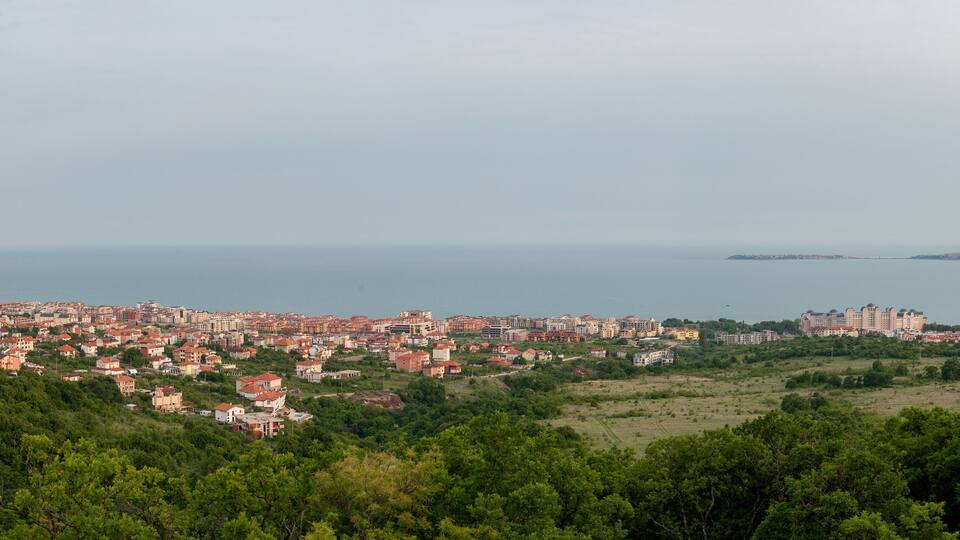 A coastal cityscape with dense housing, a large body of water, and a hilly foreground.