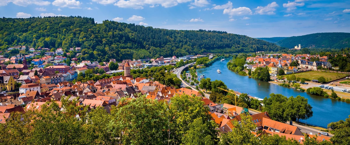 A river flowing through a town with red-roofed buildings, surrounded by lush green hills and a clear blue sky.