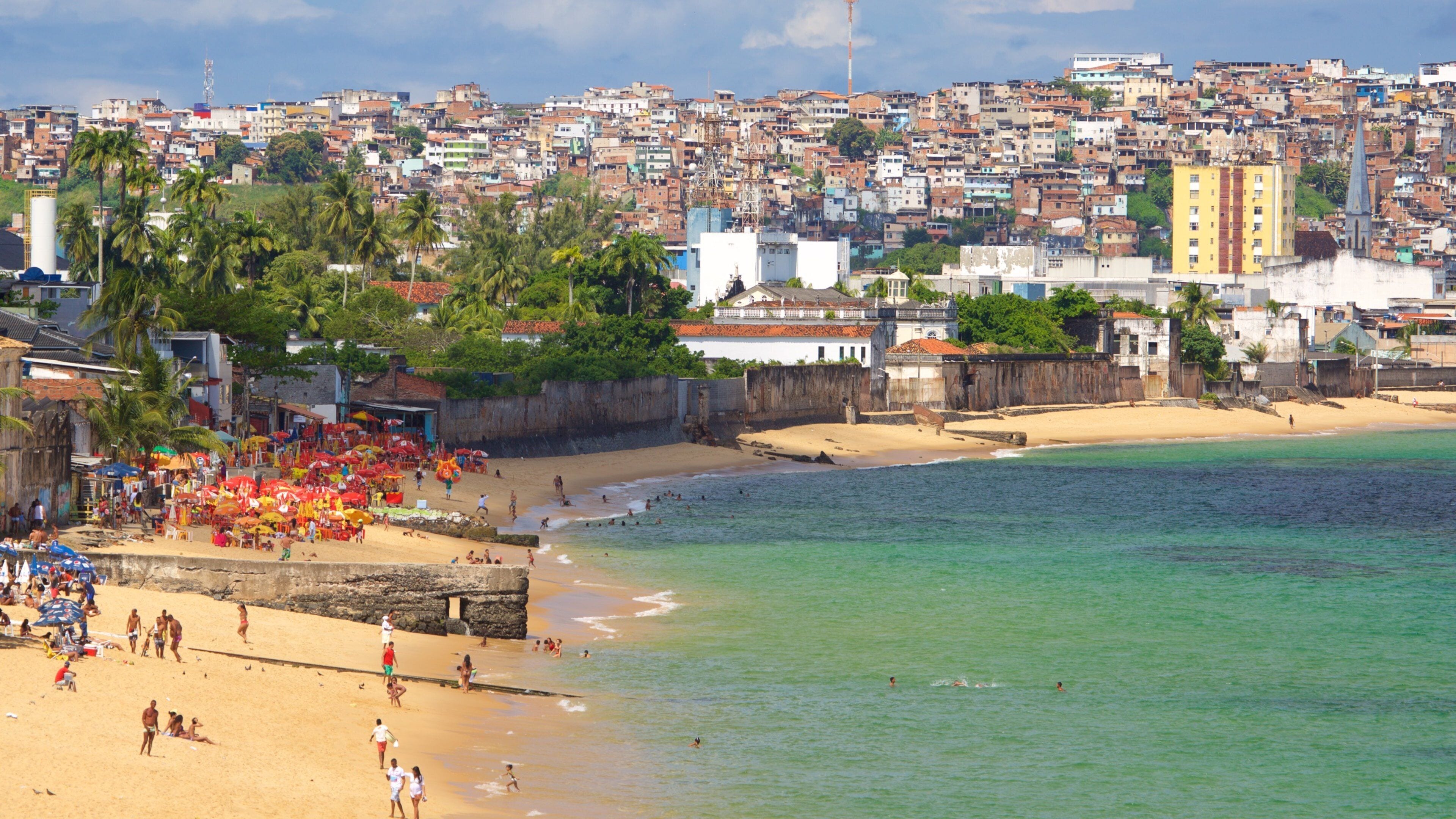 Una playa con gente, palmeras y un paisaje urbano al fondo.