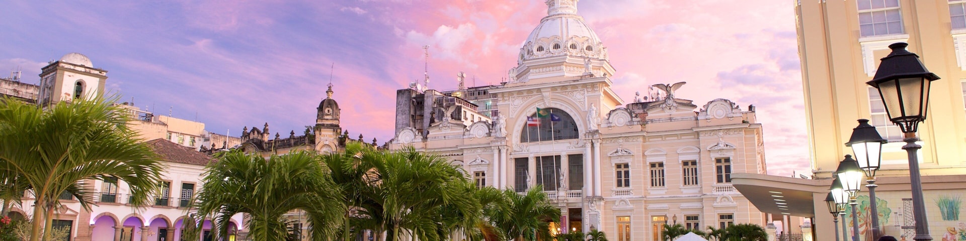 A plaza with palm trees, a historic building with a dome, and people sitting at tables.