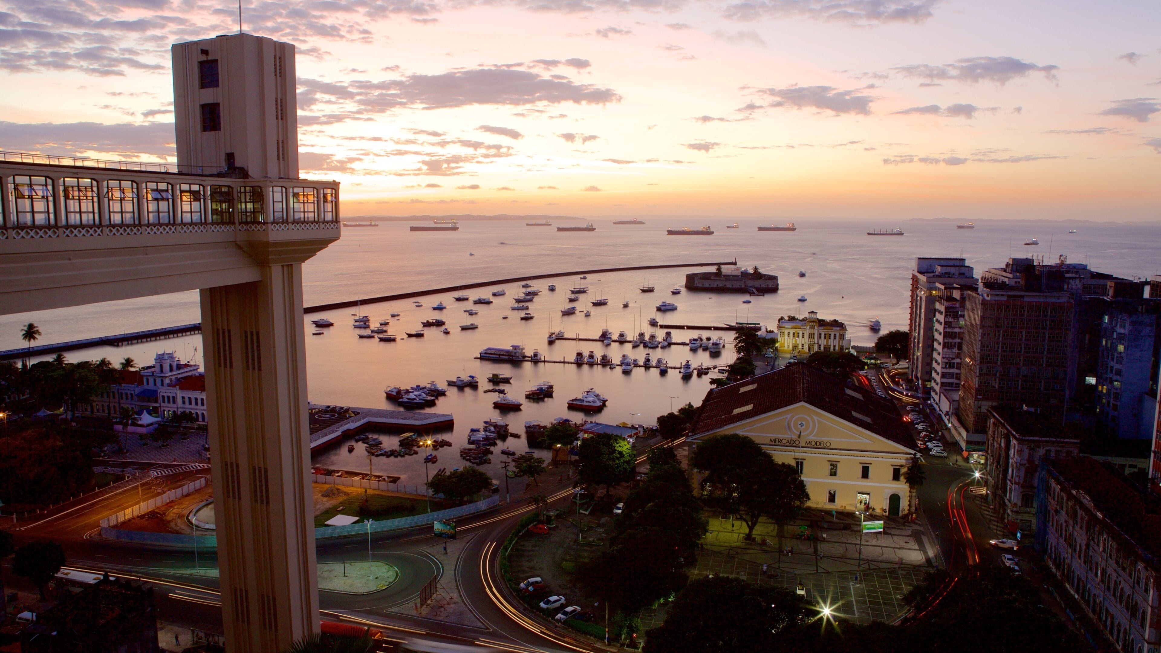 Paisagem costeira ao pôr do sol com um porto, barcos e uma ponte.