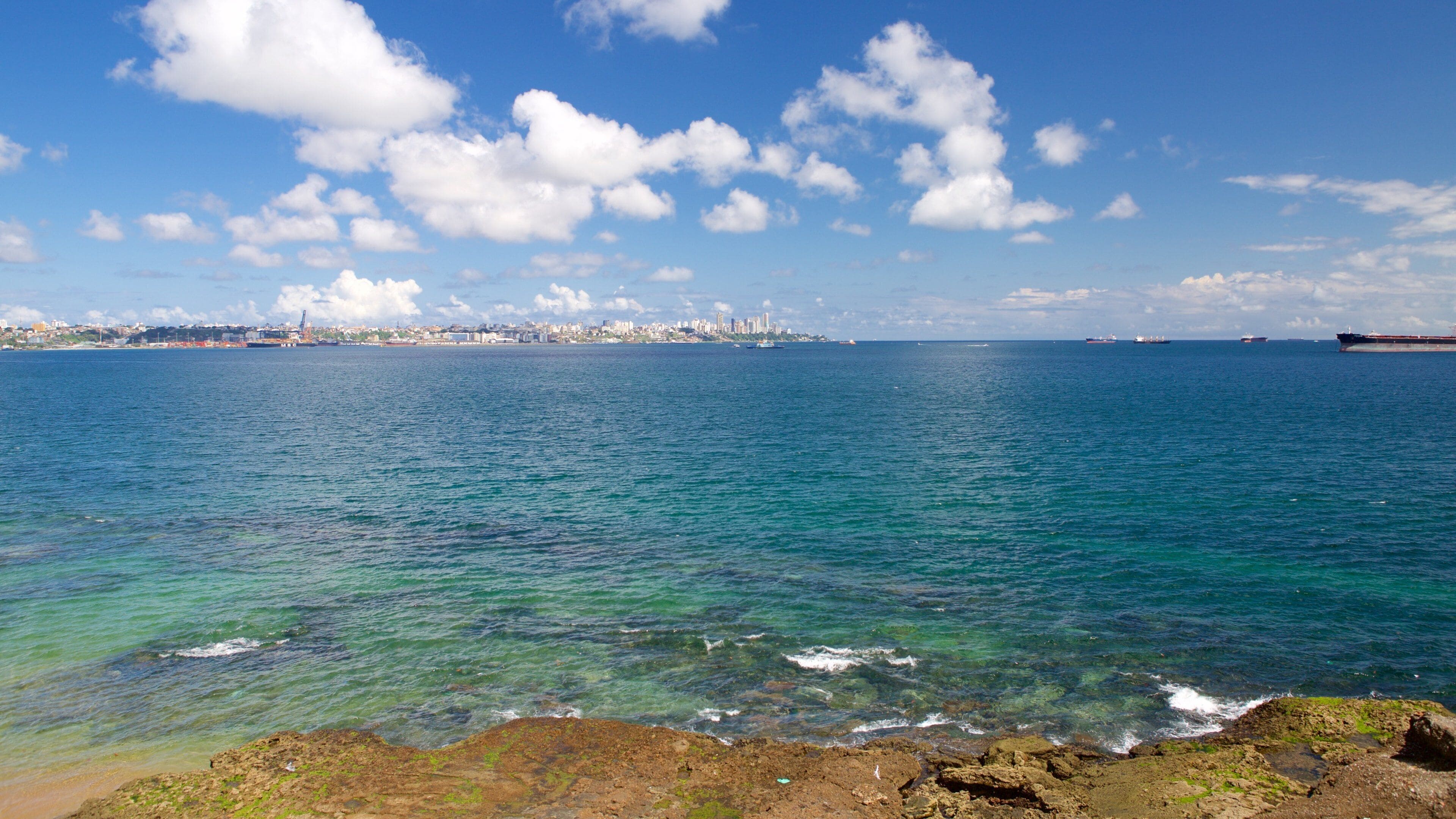 Una ciudad costera con un mar azul cristalino, un litoral rocoso y un horizonte urbano lejano bajo un cielo parcialmente nublado.