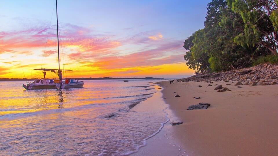 A beach with a sailboat anchored near the shore, surrounded by calm waters and a sunset sky.