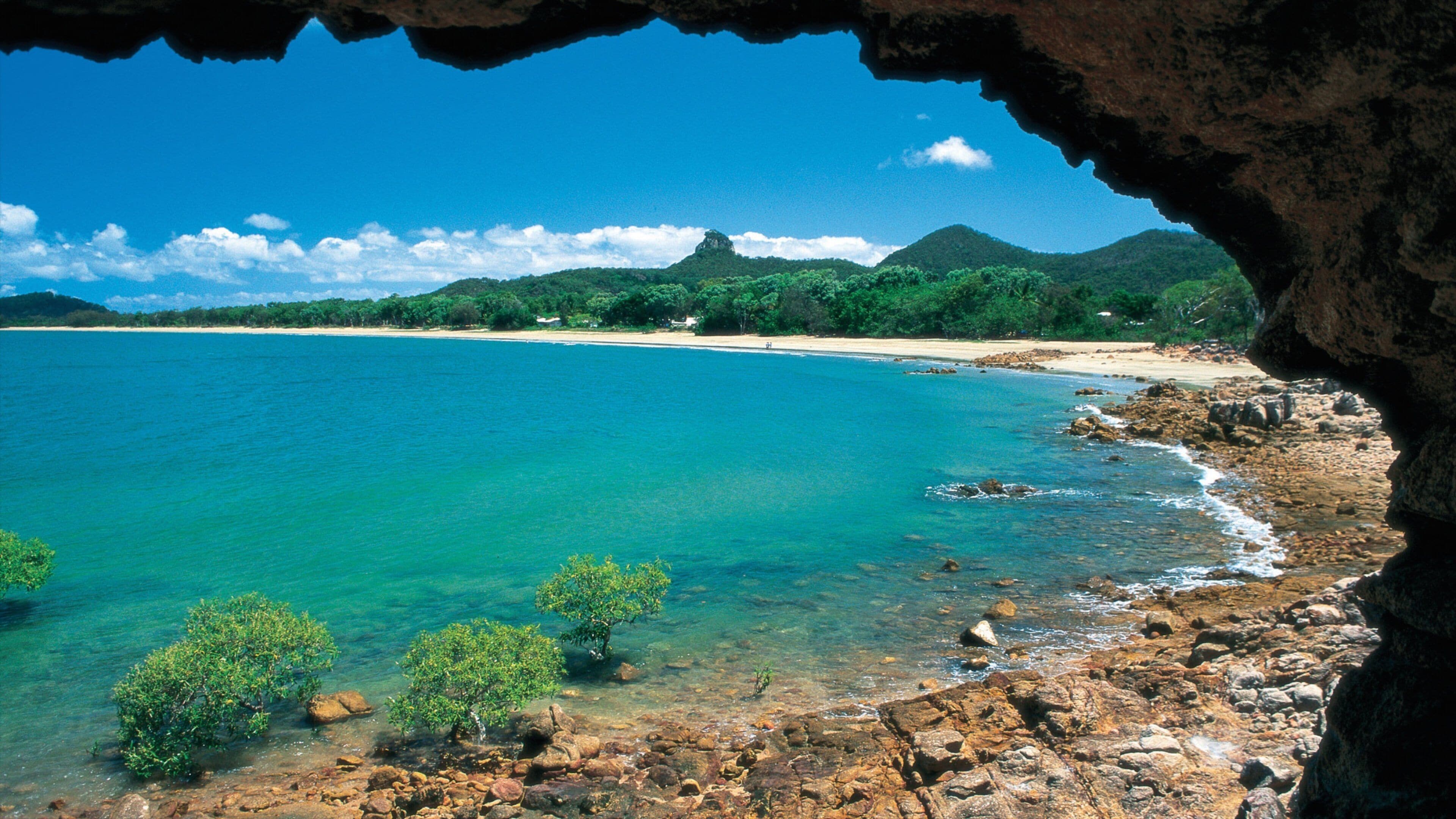 A coastal landscape with clear turquoise waters, rocky shores, and lush green hills in the background.