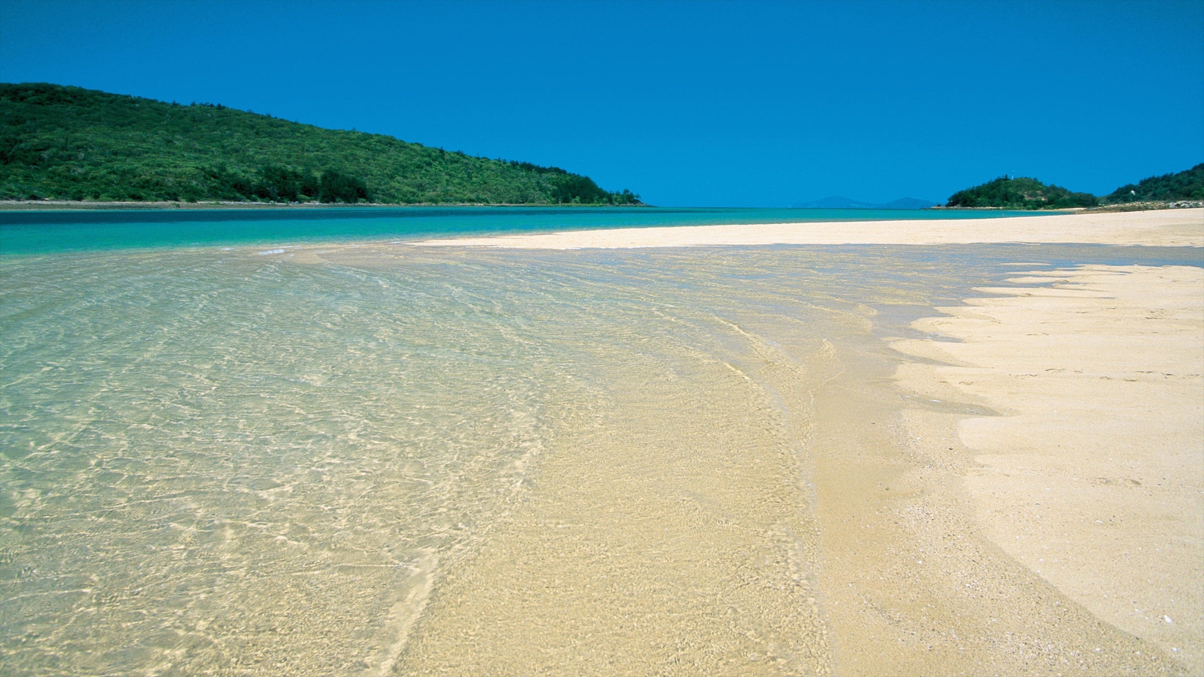 Um mar azul-turquesa translúcido, uma praia de areia e colinas verdes ao fundo.