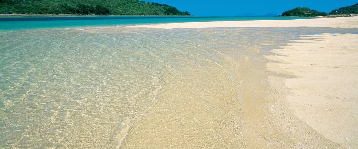 A clear turquoise sea, a sandy beach, and green hills in the background.