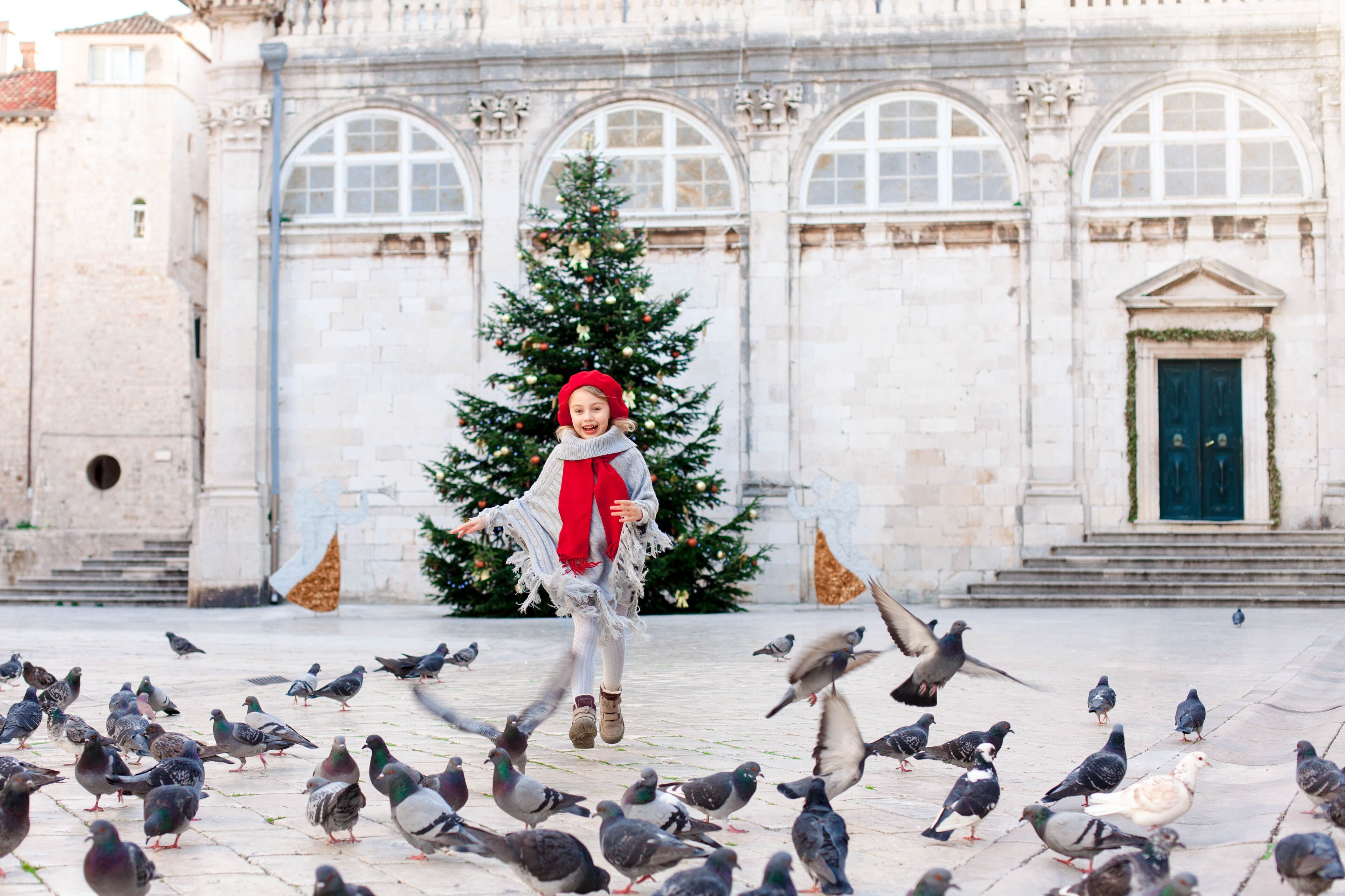 Happy kid girl is running with flying doves on square and Christmas tree in Europe. Cute child chases pigeons. Romantic vintage atmosphere in ancient town. Authentic architecture in Dubrovnik, Croatia