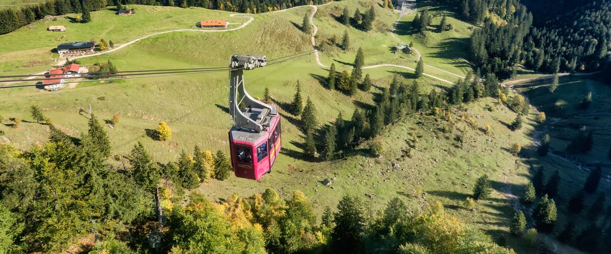 A cable car ascending a mountain with a view of a valley, forested hills, and a small settlement.