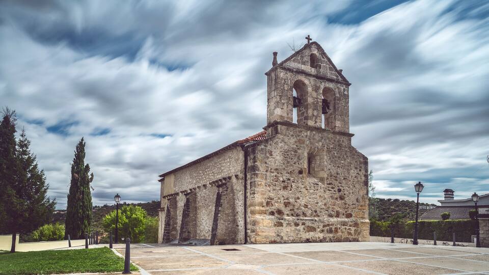 An old stone church with a bell tower and a cross on top, surrounded by a paved plaza and trees.