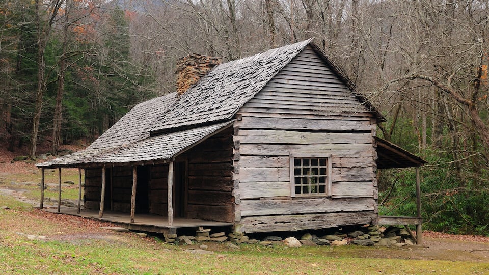 A rustic wooden cabin with a sloped roof, surrounded by a forested area with bare trees.