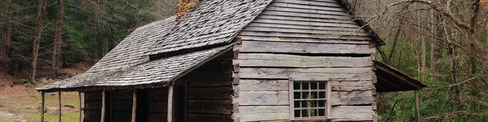 A rustic wooden cabin with a sloped roof, surrounded by a forested area with bare trees.