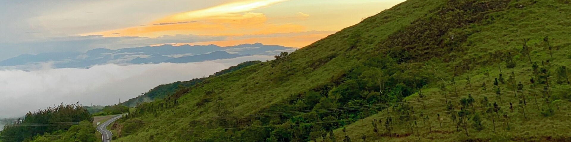 Uma estrada sinuosa serpenteia por uma encosta verdejante, com o pôr do sol ao fundo.