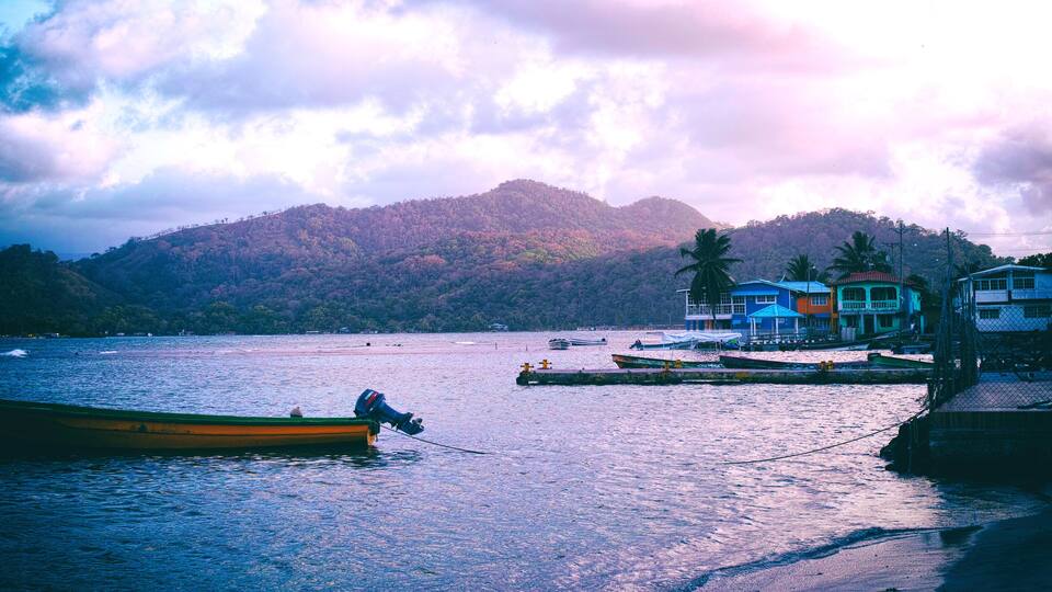 A calm lake with a small boat, a dock with colorful buildings, and a forested hill in the background.