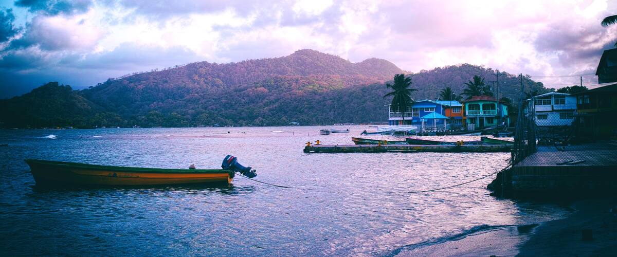 A calm lake with a small boat, a dock with colorful buildings, and a forested hill in the background.
