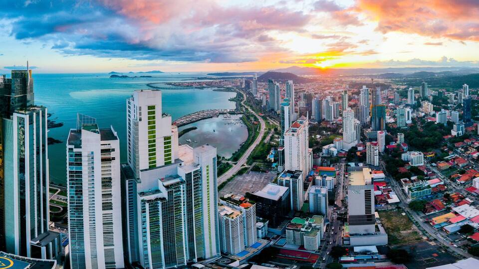 A panoramic cityscape with high-rise buildings, a waterfront, and a sunset over the horizon.
