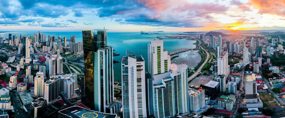 A panoramic cityscape with high-rise buildings, a waterfront, and a sunset over the horizon.
