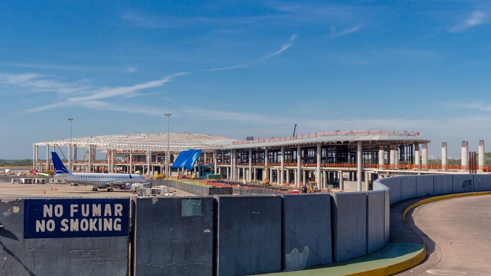 An airport terminal with a 'No Fumar No Smoking' sign, a parked airplane, and construction barriers.