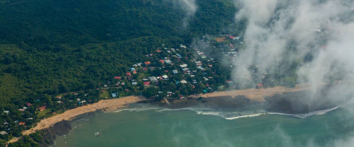 An aerial view of a coastal town with dense greenery, a beach, and a rocky shoreline.