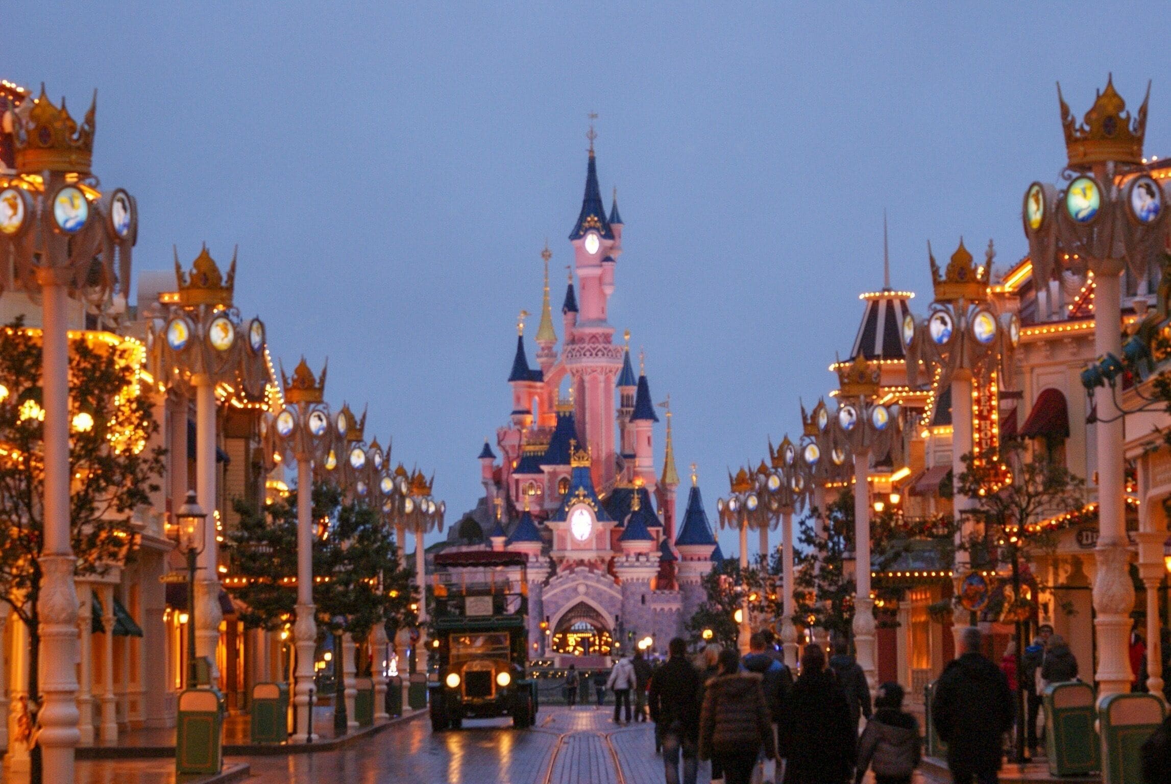 A castle-like structure with multiple spires and clocks, surrounded by well-lit columns and trees.