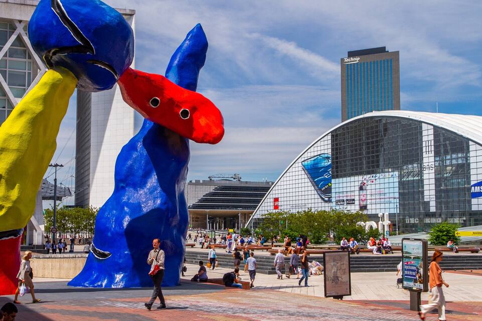 Una piazza pubblica con una grande scultura a forma di pesce, un edificio moderno con pubblicità, e persone che passeggiano.