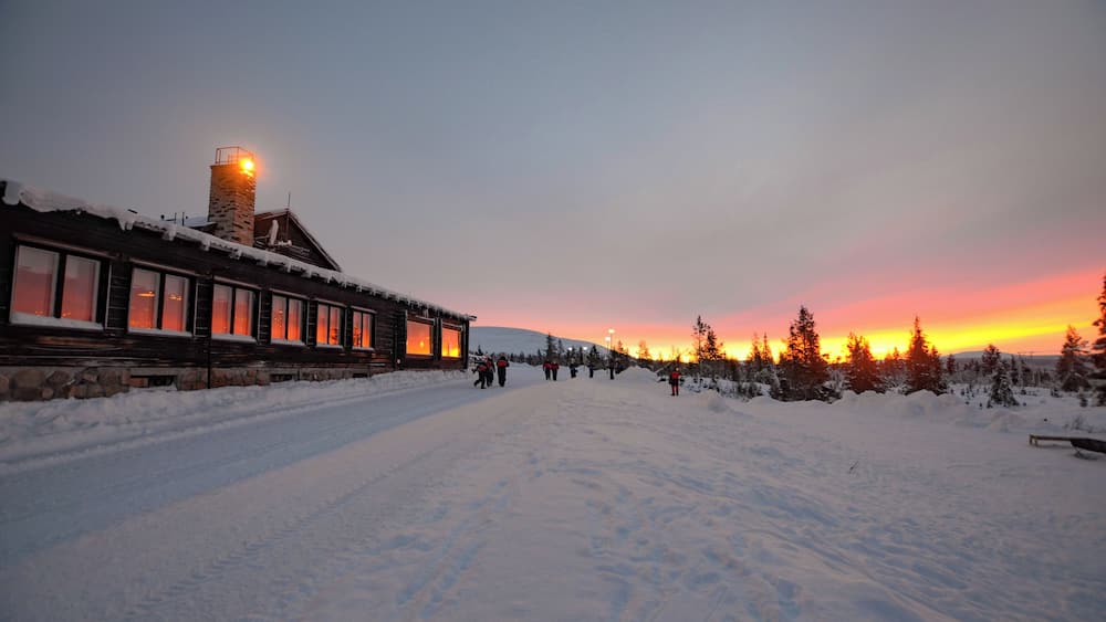 A snow-covered path leading to a lodge with a stone chimney, surrounded by evergreen trees and a sunset in the background.