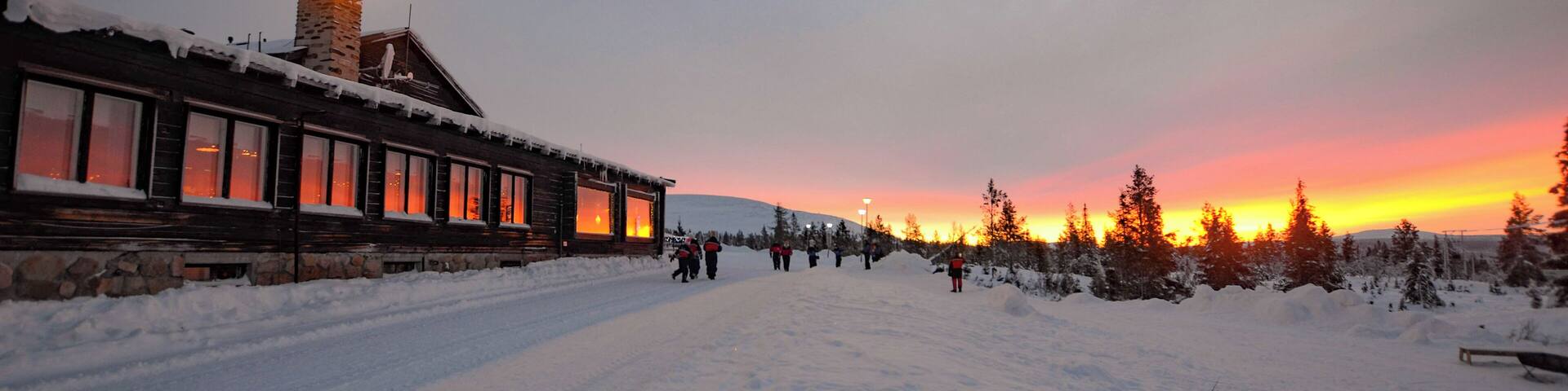 A snow-covered path leading to a lodge with a stone chimney, surrounded by evergreen trees and a sunset in the background.